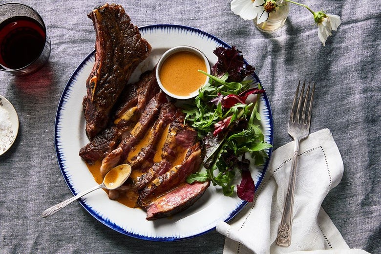 Pieces of brown and red steak soaked in orange sauce sit on a plate with greens, next to a fork on a napkin, a beverage and a flower in a vase.